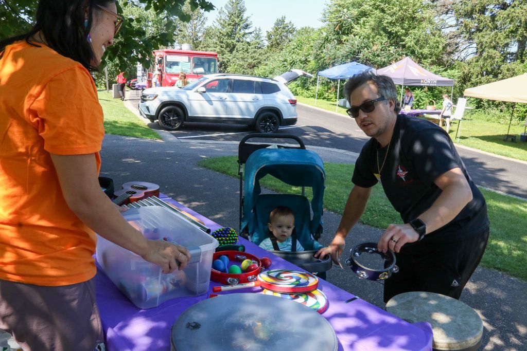Two people at a table with musical instruments, a baby in a stroller, and a car parked in the background.