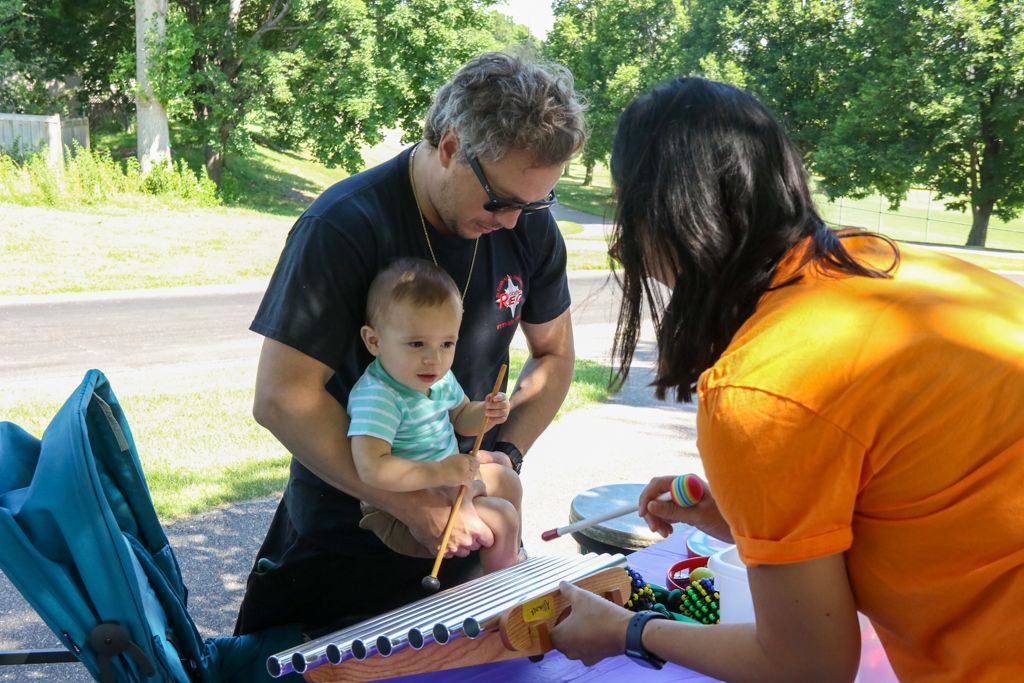 Man holding baby points to a weaving loom as a woman watches. Outdoors, sunny day.