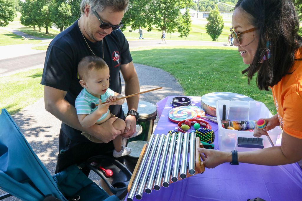 Man holds baby near a table with musical instruments. Woman offers instrument to baby. Outdoors setting.