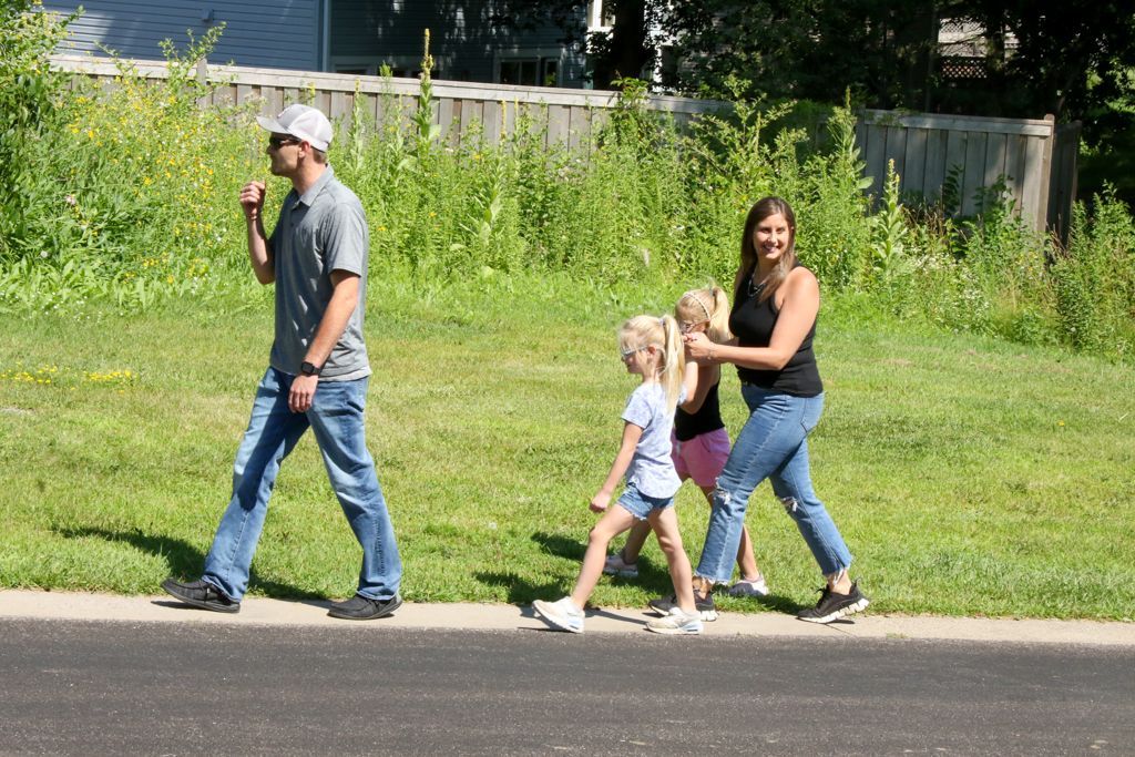 A family of four walking on a sidewalk. The father gestures with his hand. The mother holds a girl's arm.