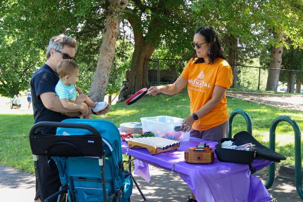Man holding baby at purple table with woman handing something to them in a park.