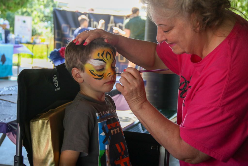 Woman paints a tiger face on a child's face at an outdoor event.