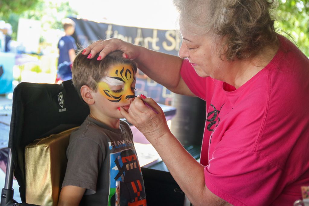 Woman painting a tiger face on a child's face outdoors. Child sits in a chair.