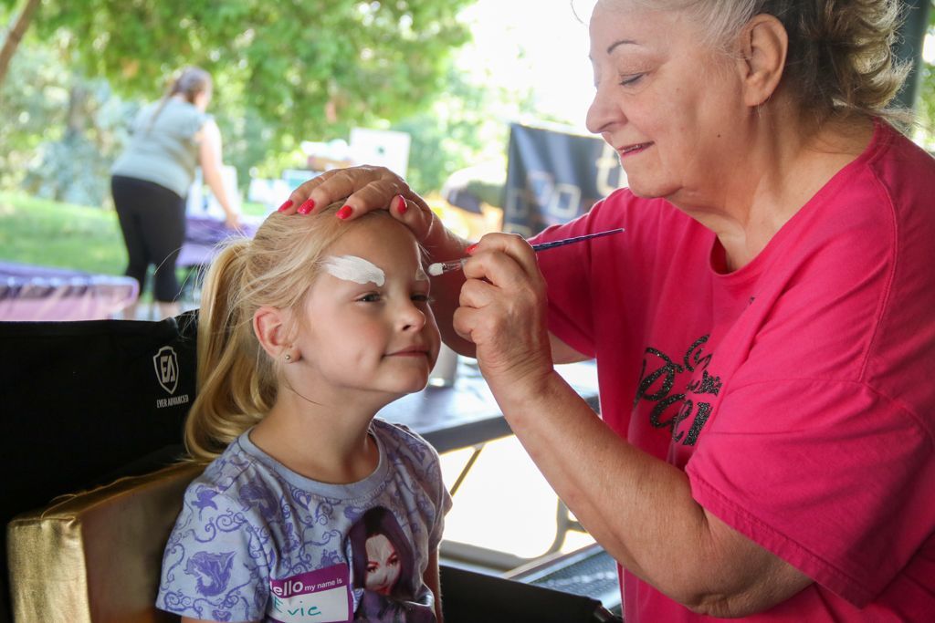 Woman painting face of girl with white eyebrows outdoors.