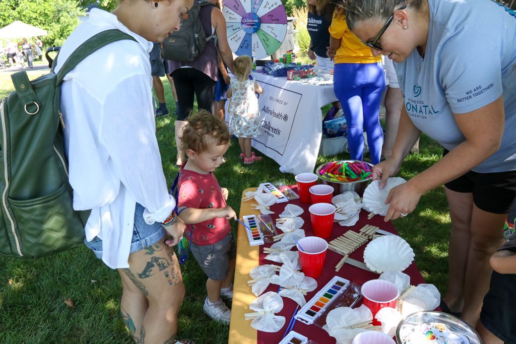 People at an outdoor craft table; a child and an adult interacting with craft supplies, other attendees visible in the background.
