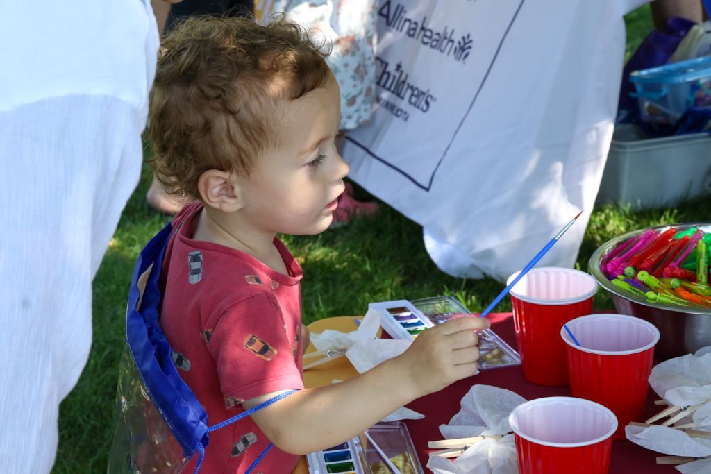 Child with curly hair painting at an outdoor table with paint and red cups.