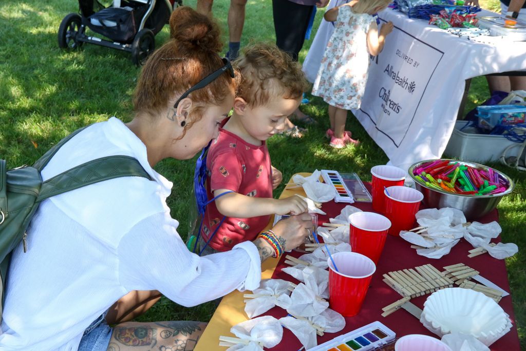 Woman and child painting at an outdoor craft table with red cups, paint, and popsicle sticks.
