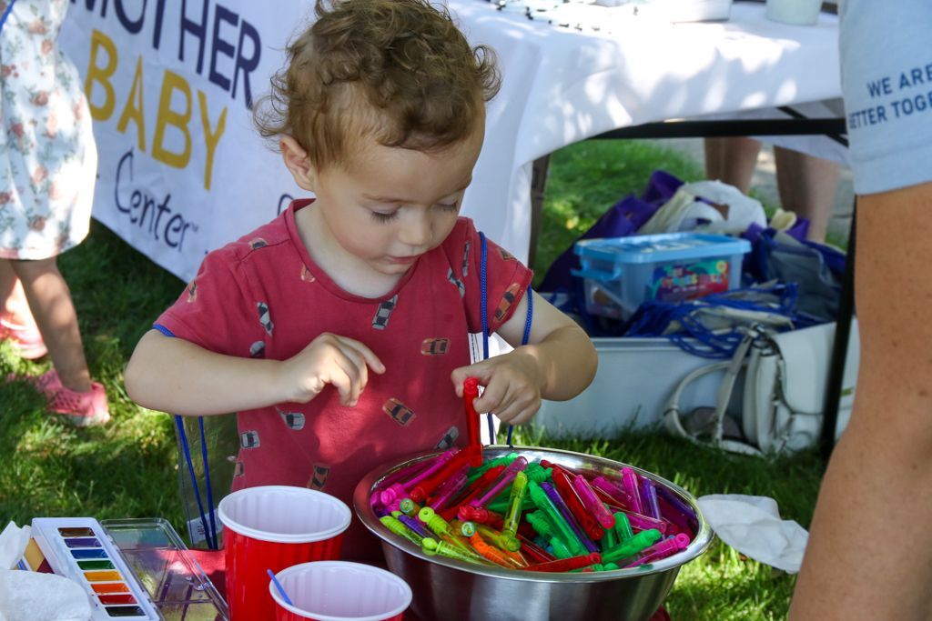 Child in red shirt selecting crayons from a bowl at a table with art supplies outdoors.