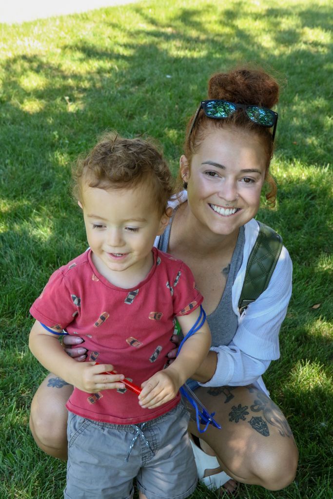 Woman smiles, holding a toddler on green grass; both have reddish-brown hair. The toddler holds a toy.