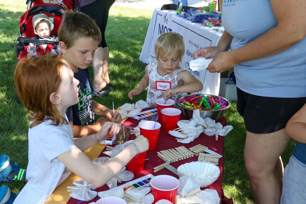 Children at an outdoor craft table, working with supplies like cups and craft sticks.