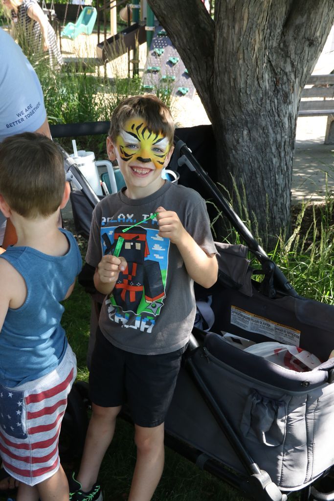 Boy with tiger face paint smiles, standing next to a stroller and another child with an American flag-print skirt.