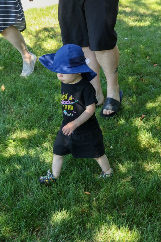 Child in blue hat and black clothes walking on grass, with adults nearby.