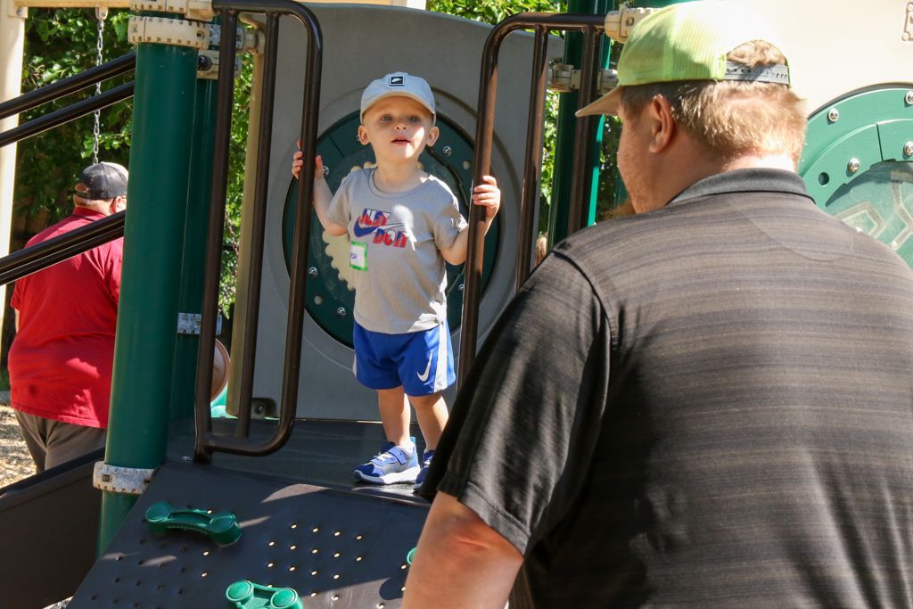 Child in a baseball cap on playground equipment, smiling, with a man watching.