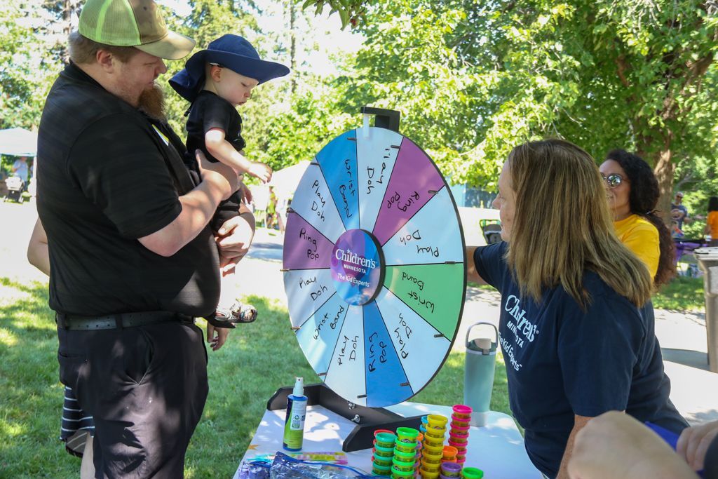 People at an outdoor event spinning a prize wheel. Man holds child, woman points at wheel.