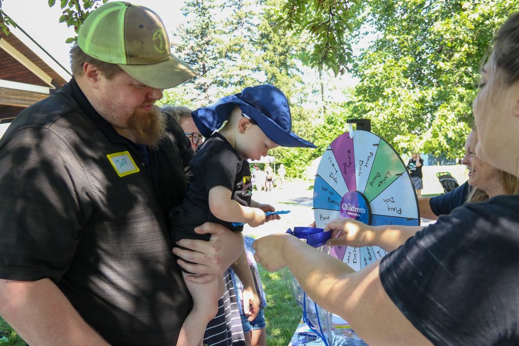 Man holding child by a prize wheel; another person spins it. Outdoors, sunny.