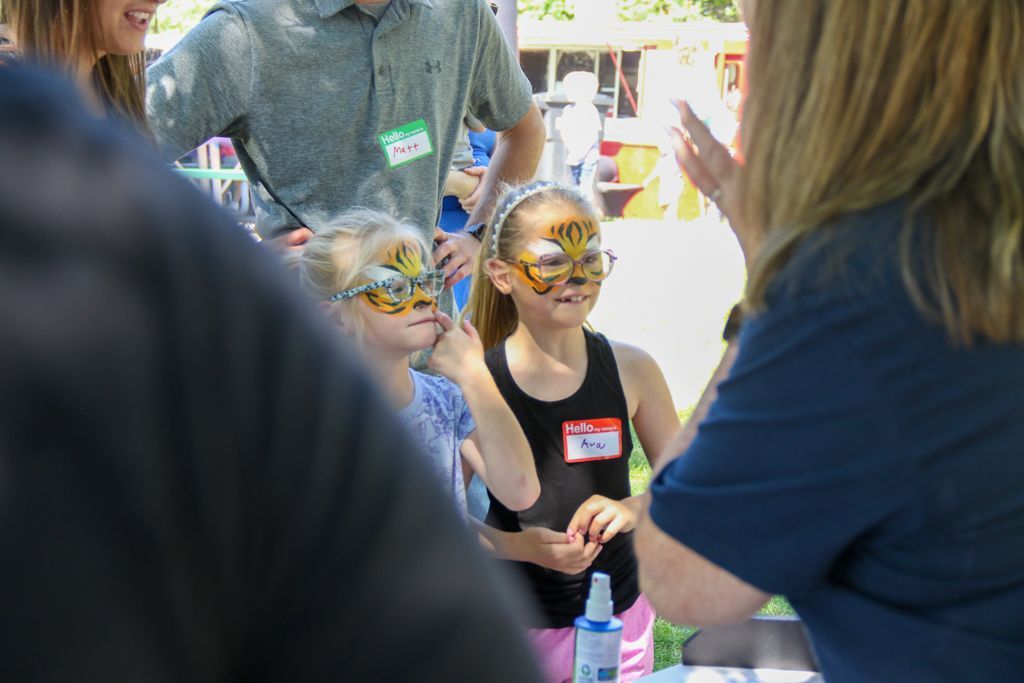 Two girls with tiger face paint pose near an adult; outdoor event.