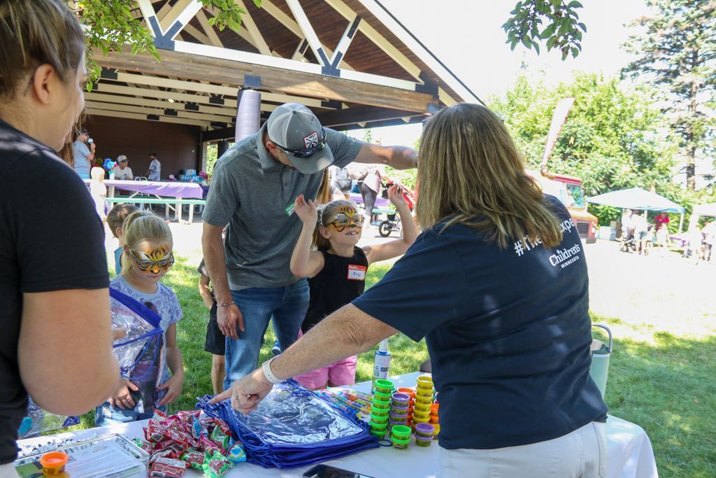 People at a table with kids and a man reaching up. A woman stands by a table with toys and crafts, outdoors.