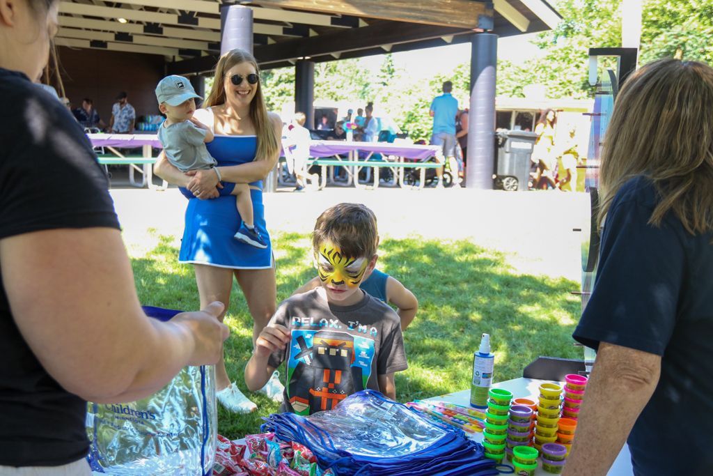 Boy with face paint at a table with toys, near woman holding a child and other people outside.
