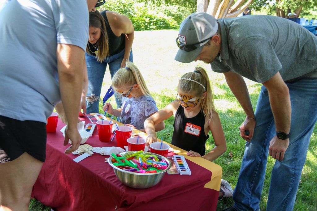 People at an outdoor craft table, two girls painting with adults looking on, surrounded by art supplies.