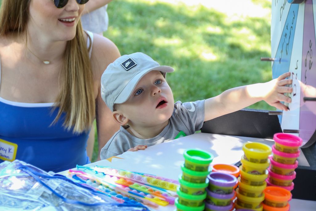 Young child reaching towards a game, next to woman, colorful playdough, outdoors.