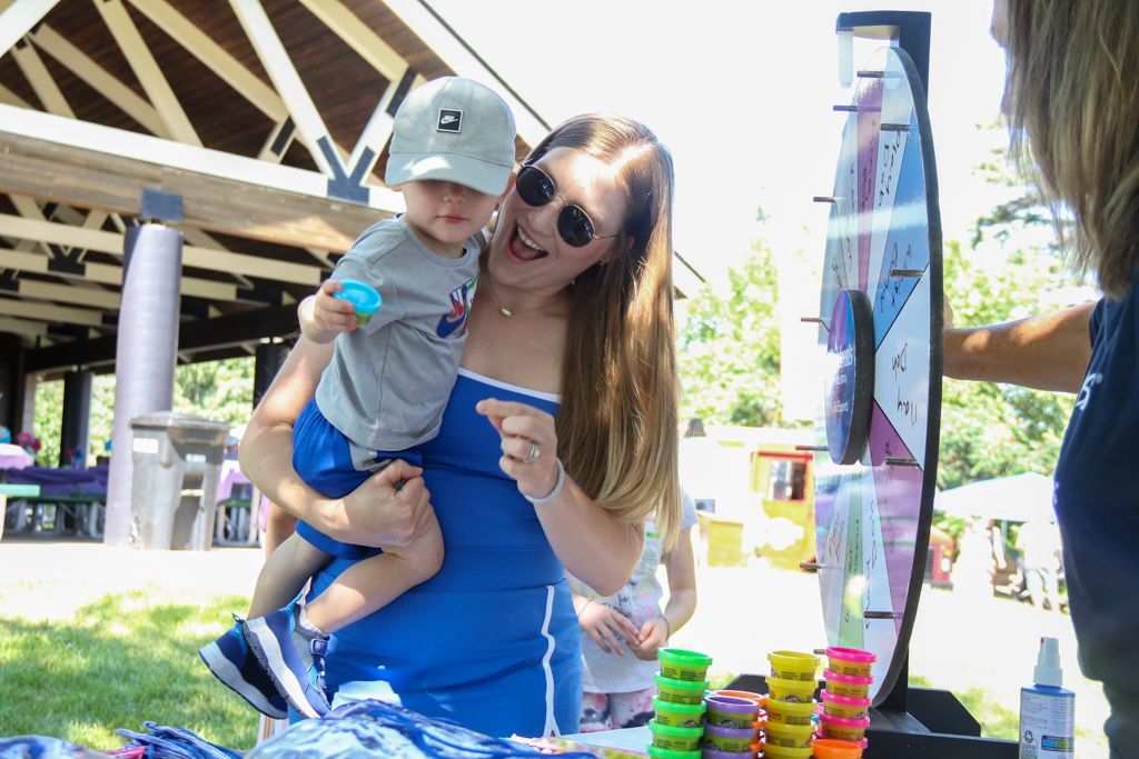 Woman holding child smiles excitedly near a prize wheel at an outdoor event.