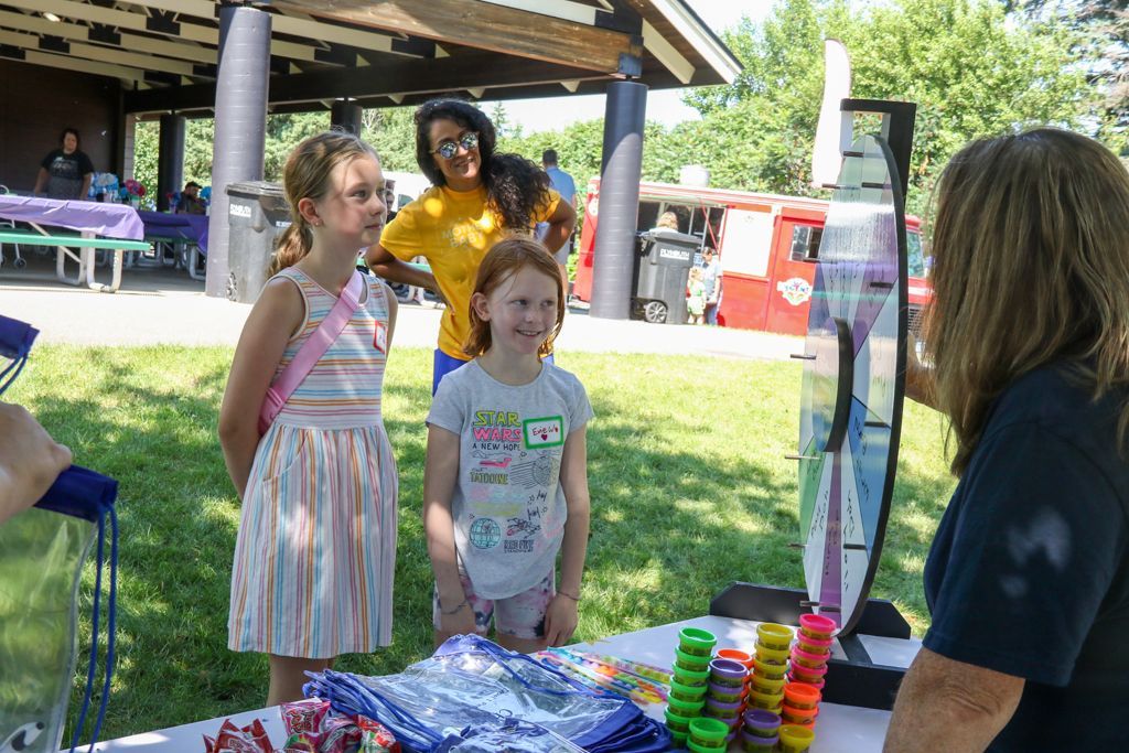 Children at a park booth, interacting with a vendor; a person in yellow watches.