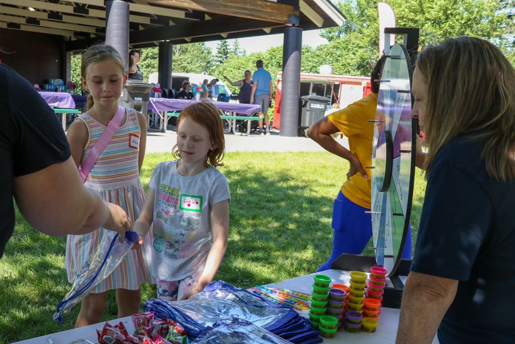 Children at a table looking at items. Two girls are focused, another person is assisting them. Outdoor setting.