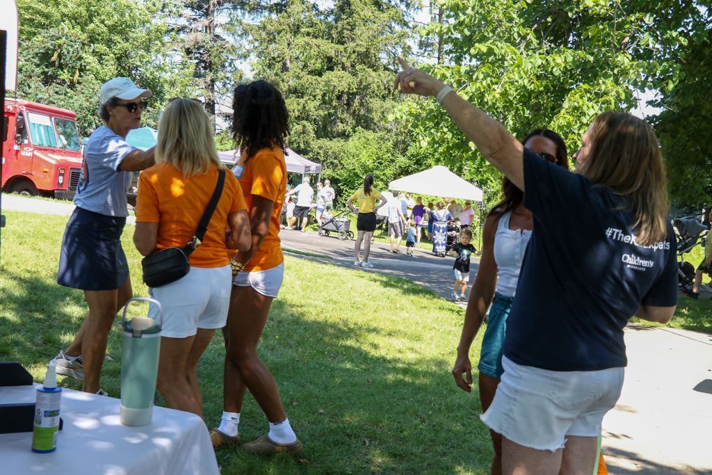 People at an outdoor event, some in orange shirts, one pointing. Others walking and a red vehicle in background.