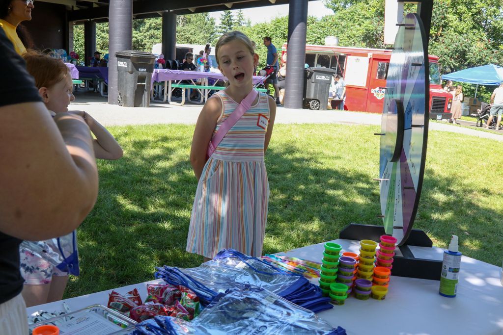 Girl with pink sash standing at a table with colorful toys, outdoors on a sunny day.