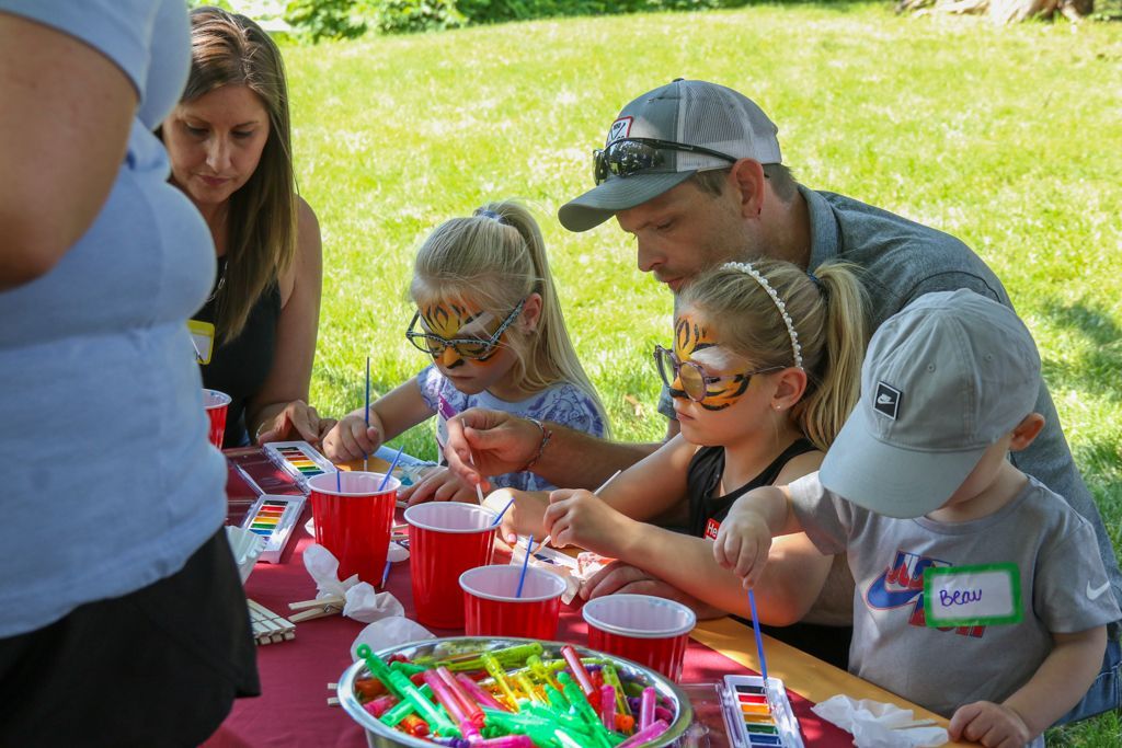 Family at a table painting with watercolors outdoors. Children have face paint on, and adults are assisting them.