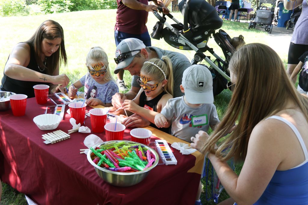 People at an outdoor table painting. Children with face paint, adults helping. Red cups, craft supplies.