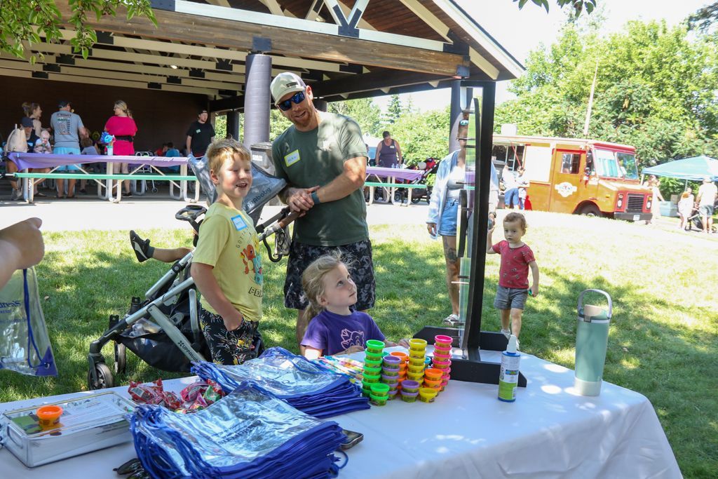 Children at a fair booth, smiling. Person behind them with bubbles, table with items. Food truck in background.