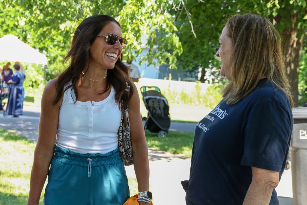 Two women smiling, chatting outdoors on a sunny day. One wears sunglasses, white top, turquoise pants. The other has on a blue t-shirt.