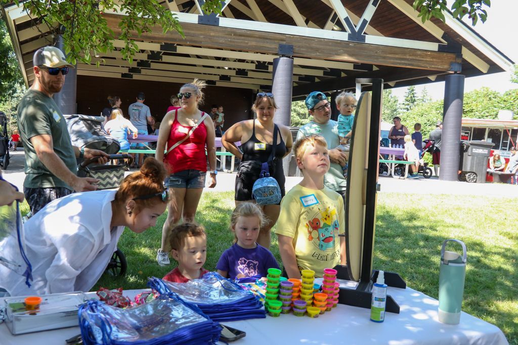People at a park booth; children looking at a mirror, colorful stacks of toys, and adults in the background.