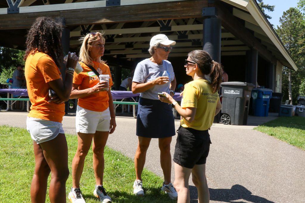 Four women conversing outside near a pavilion. Three wear orange shirts, one yellow. Outdoors on a sunny day.
