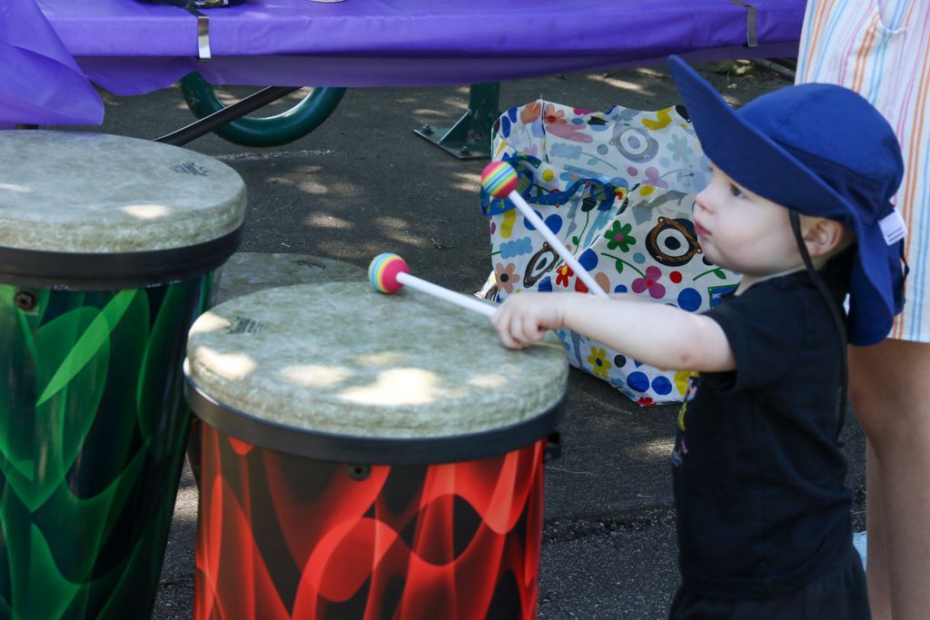 Young child in blue hat playing a colorful drum outside.