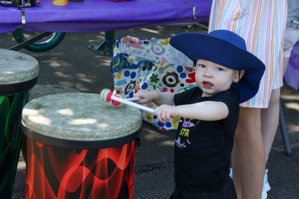Young child wearing blue hat plays drum outdoors.