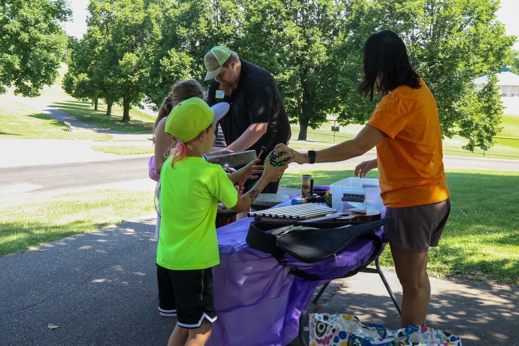 People at a table. A man in a hat and a woman in orange hand items to children at an outdoor event.