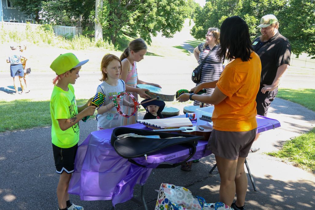 Children at a table with an adult; selecting items. Outdoor setting with trees and sidewalk.