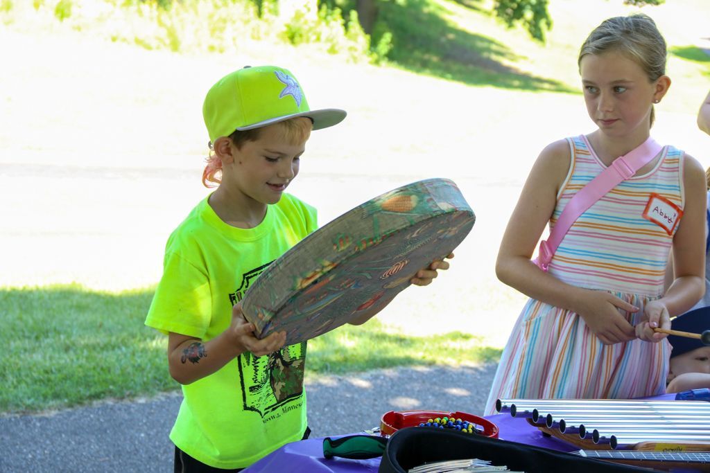 Boy in neon shirt and hat looking at a musical instrument; girl in striped dress watches.