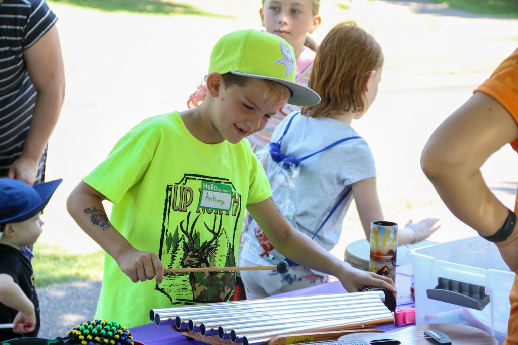 Boy playing a xylophone outdoors, wearing a neon green shirt and hat. Others watch.