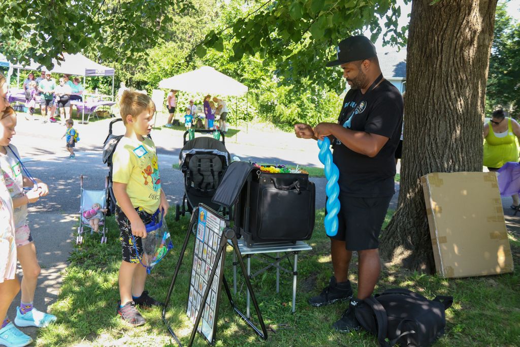 Person twisting a blue balloon, interacting with a child at an outdoor event, with trees and a vendor stall in the background.