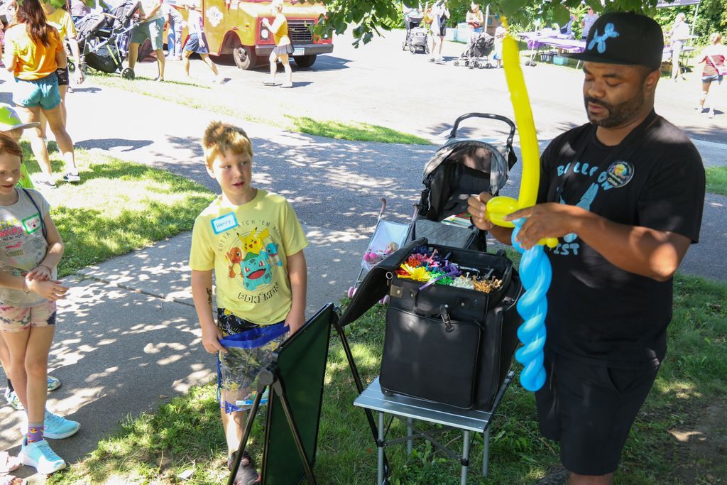 Man making balloon animal for a boy at outdoor event; black bag, table.