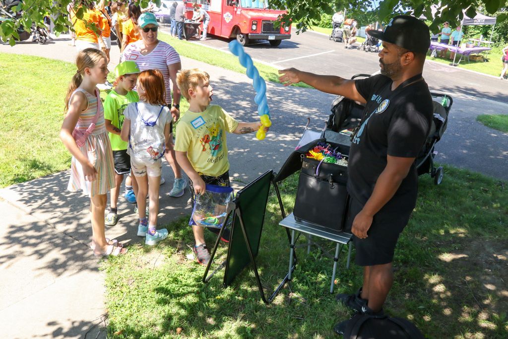 Balloon artist giving a sword to a child, surrounded by other children on a sunny day.