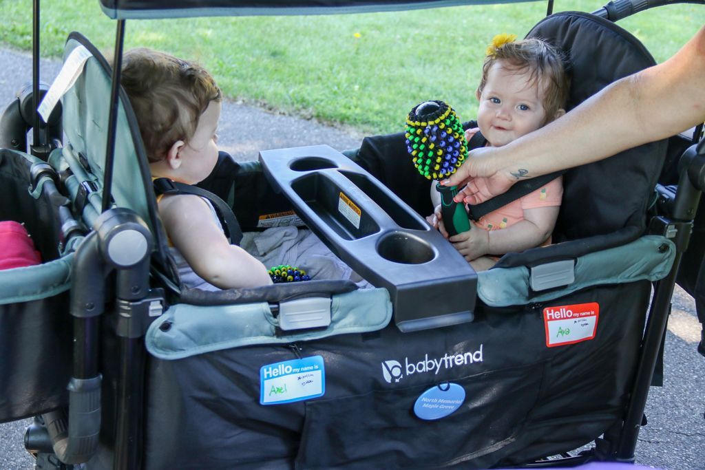Two babies sit in a Baby Trend wagon. One plays with a rattle, the other looks away. A person's hand reaches in.