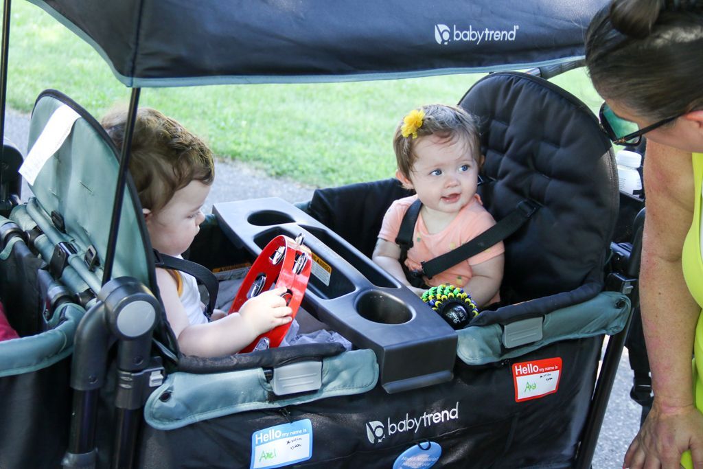 Two babies in a wagon with a canopy. One plays a tambourine. Green wagon with black accents.