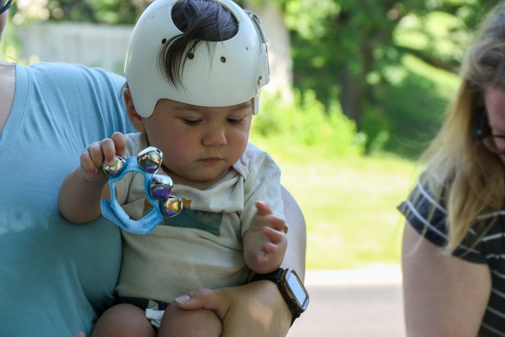 Baby in a white helmet, holding a rattle, being held by an adult; sunny outdoor setting.