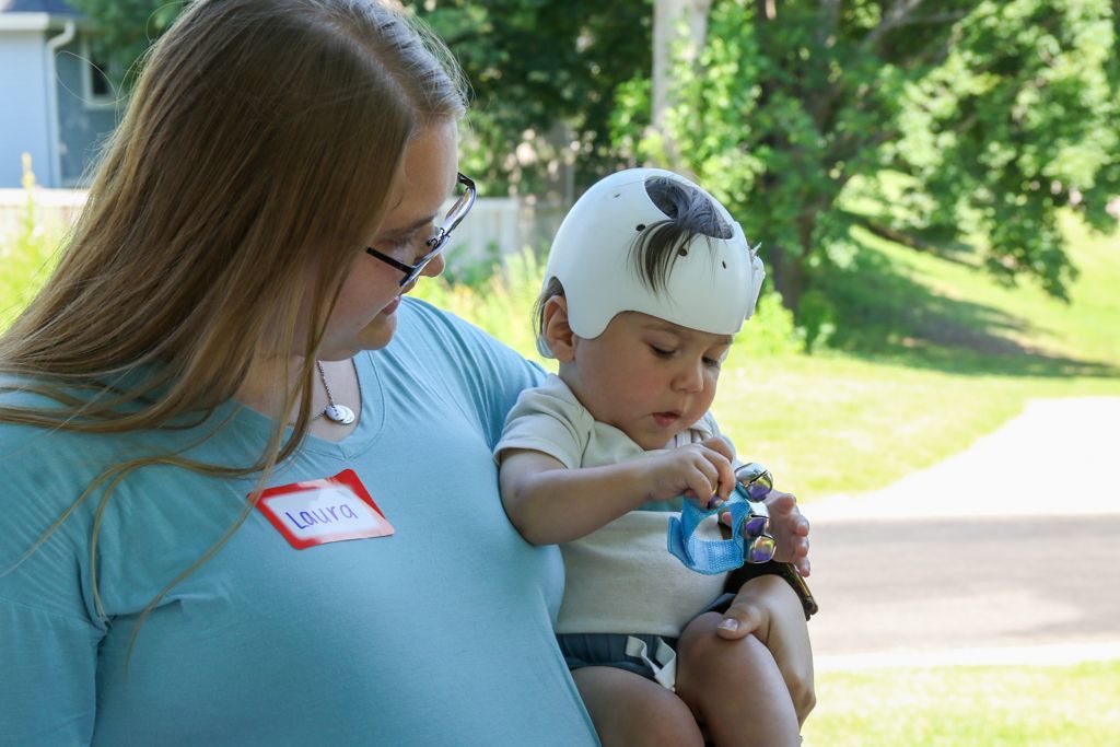 Woman holding baby in a helmet outdoors, baby plays with a toy.