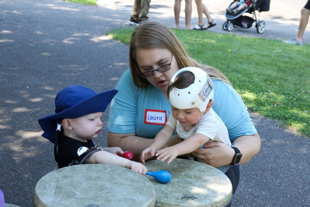Woman with two children playing drums outdoors; one child wears a helmet.