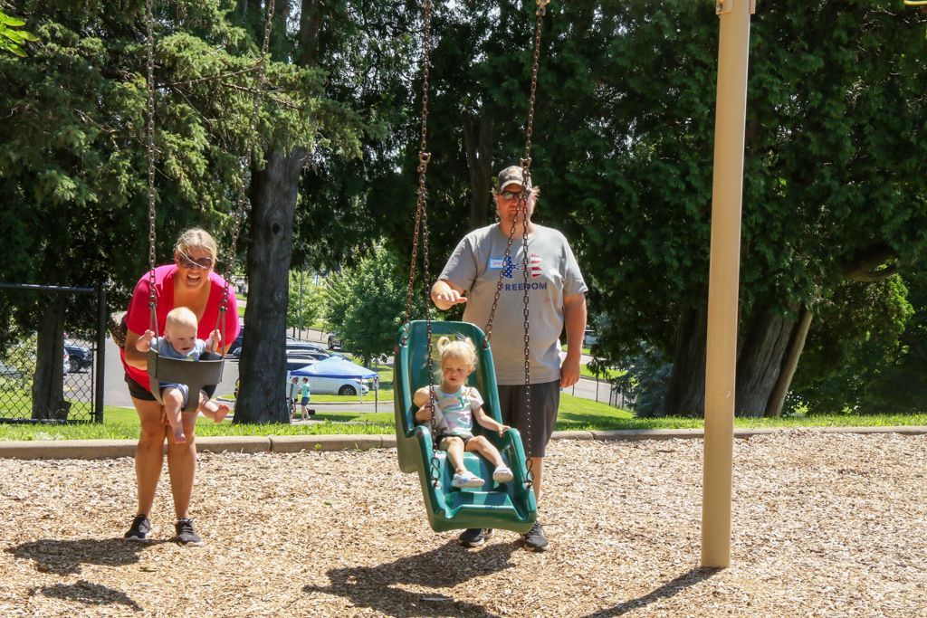 People at a playground: A woman holds a baby, a man stands by a swing with a toddler in it, mulch ground.